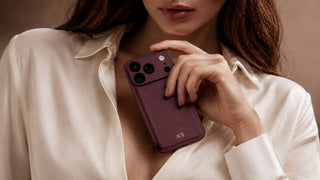 Woman in a cream silk shirt holding a burgundy personalised leather iPhone case with gold initials