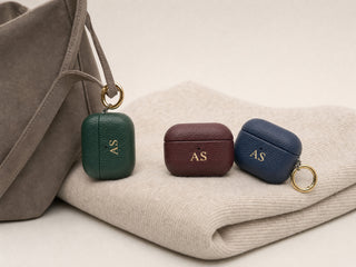 Personalised leather AirPods cases in green, burgundy and navy with gold rings on a cashmere sweater