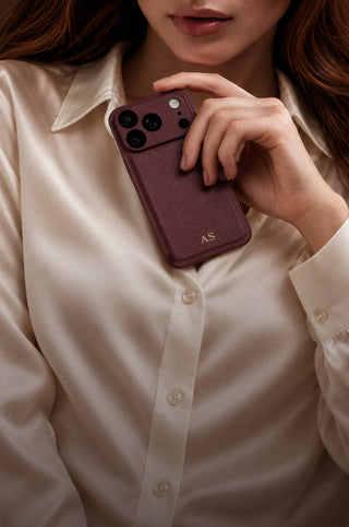 Burgundy vegan leather phone case with gold initials held against a cream silk blouse